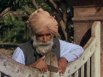 Movie still from “The River” (1951), directed by Jean Renoir – An old man with a beard and a turban is leaning on a fence; Medium shot, Low angle