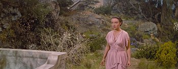 Movie still from “The Robe” (1953), directed by Henry Koster – A woman in a pink dress standing next to a tree stump; Medium shot, Over the shoulder angle
