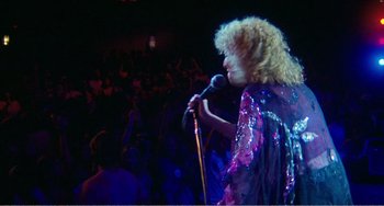 Movie still from “The Rose” (1979), directed by Mark Rydell – A woman in a purple dress holding a microphone in front of an audience; Medium shot, Low angle