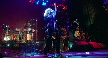 Movie still from “The Rose” (1979), directed by Mark Rydell – A man with a microphone on a stage with a group of people; Wide shot, Low angle