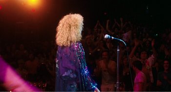 Movie still from “The Rose” (1979), directed by Mark Rydell – A woman in a purple jacket is on a stage with a microphone; Wide shot, Over the shoulder angle