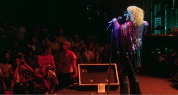 Movie still from “The Rose” (1979), directed by Mark Rydell – A man with a microphone on a stage with people watching; Wide shot, Low angle
