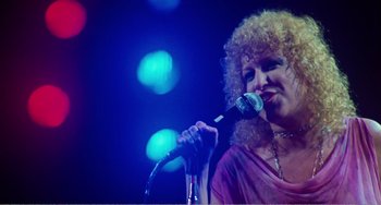 Movie still from “The Rose” (1979), directed by Mark Rydell – A woman with curly blonde hair is singing into a microphone; Medium shot, Low angle