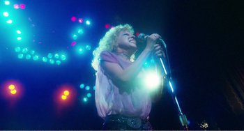 Movie still from “The Rose” (1979), directed by Mark Rydell – A woman with curly hair is singing into a microphone; Wide shot, Low angle