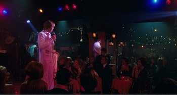 Movie still from “The Rose” (1979), directed by Mark Rydell – A woman in a white suit is holding a microphone in front of a group of people; Wide shot, Over the shoulder angle