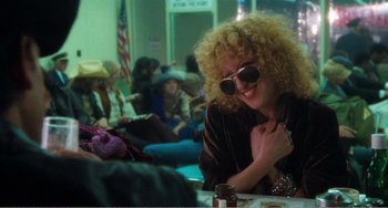 Movie still from “The Rose” (1979), directed by Mark Rydell – A woman sitting at a table in a room with other people; Medium shot, Over the shoulder angle