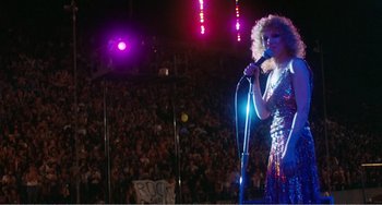 Movie still from “The Rose” (1979), directed by Mark Rydell – A woman in a sequined dress is holding a microphone; Wide shot, Low angle