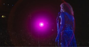 Movie still from “The Rose” (1979), directed by Mark Rydell – A woman in a blue dress holding a microphone on a stage; Wide shot, Over the shoulder angle