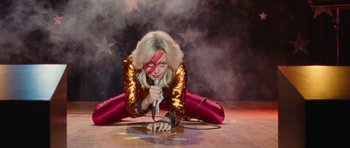 Movie still from “The Runaways” (2010), directed by Floria Sigismondi – A woman in a red and gold outfit holding a microphone; Wide shot, Overhead angle