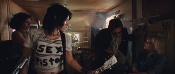 Movie still from “The Runaways” (2010), directed by Floria Sigismondi – A woman holding a guitar in a room with other people; Medium shot, Low angle