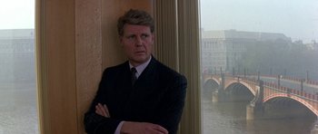 Movie still from “The Russia House” (1990), directed by Fred Schepisi – A man in a suit leaning against a wall with his arms crossed; Medium shot, Over the shoulder angle