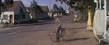 Movie still from “The Russians Are Coming the Russians Are Coming” (1966), directed by Norman Jewison – A man riding a bike down a street; Wide shot, Over the shoulder angle