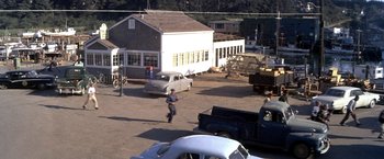 Movie still from “The Russians Are Coming the Russians Are Coming” (1966), directed by Norman Jewison – A man walking across a dirt road in front of a building; Extreme Wide shot, High angle
