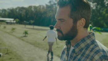 Movie still from “The Sacrament” (2013), directed by Ti West – A man with a goatee looks to his left while a boy in a white t - shirt rides a skate board behind him; Close Up shot, Over the shoulder angle