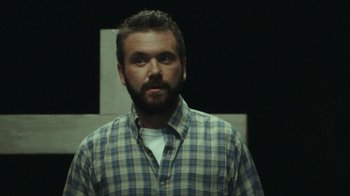 Movie still from “The Sacrament” (2013), directed by Ti West – A man in a plaid shirt standing in front of a cross; Close Up shot, Low angle