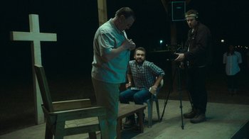 Movie still from “The Sacrament” (2013), directed by Ti West – Two men and a man sitting on a bench in front of a camera; Medium shot, Over the shoulder angle
