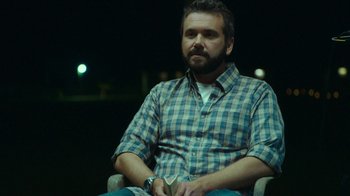 Movie still from “The Sacrament” (2013), directed by Ti West – A man sitting in a chair in a dark room; Medium shot, Over the shoulder angle