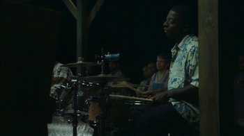 Movie still from “The Sacrament” (2013), directed by Ti West – A man playing drums in front of a group of people; Medium shot, Over the shoulder angle