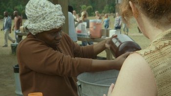 Movie still from “The Sacrament” (2013), directed by Ti West – A man wearing a white hat and a brown shirt is working on something in a metal bucket; Medium shot, Over the shoulder angle