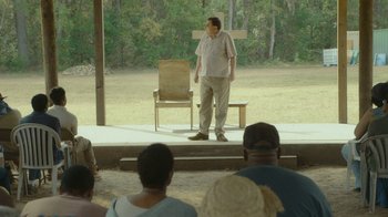 Movie still from “The Sacrament” (2013), directed by Ti West – A man standing on a stage in front of a crowd; Wide shot, Over the shoulder angle