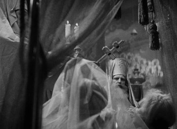 Movie still from “The Scarlet Empress” (1934), directed by Josef von Sternberg – An old man dressed as a priest holding a crucifix; Medium shot, Low angle
