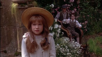 Movie still from “The Secret Garden” (1993), directed by Agnieszka Holland – A young girl wearing a straw hat in front of two men sitting on a bench in a flower garden; Medium shot, Over the shoulder angle