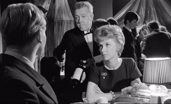 Movie still from “The Servant” (1963), directed by Joseph Losey – An older man and a woman at a table with a bottle of wine in front of them; Medium shot, Over the shoulder angle