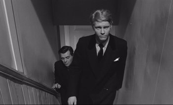 Movie still from “The Servant” (1963), directed by Joseph Losey – A man in a suit and tie standing next to another man in a suit and tie; Medium shot, Low angle