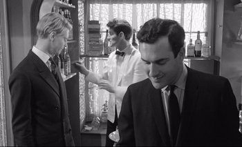 Movie still from “The Servant” (1963), directed by Joseph Losey – A man in a suit and a bow tie is standing next to another man in a suit and a tie; Medium shot, Over the shoulder angle