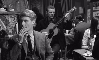 Movie still from “The Servant” (1963), directed by Joseph Losey – A man smoking a cigarette while a man plays a guitar; Medium shot, Over the shoulder angle