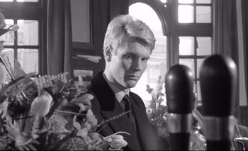 Movie still from “The Servant” (1963), directed by Joseph Losey – Black and white photograph of a man in a suit and tie in front of a microphone; Close Up shot, Low angle