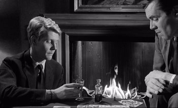 Movie still from “The Servant” (1963), directed by Joseph Losey – A man sitting at a table with a glass of wine in front of a fireplace; Medium shot, High angle