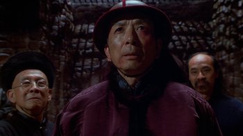 Movie still from “The Shadow” (1994), directed by Russell Mulcahy – An older man wearing a red hat and a purple jacket; Close Up shot, Over the shoulder angle