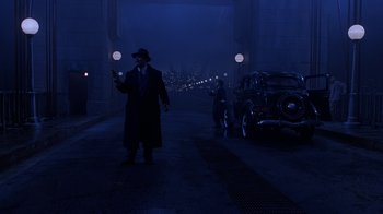Movie still from “The Shadow” (1994), directed by Russell Mulcahy – A man in a black coat and a hat is holding a flashlight; Wide shot, Over the shoulder angle
