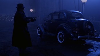 Movie still from “The Shadow” (1994), directed by Russell Mulcahy – A man holding a gun standing next to a car in the dark; Wide shot, Over the shoulder angle