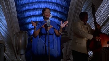 Movie still from “The Shadow” (1994), directed by Russell Mulcahy – A woman in a blue dress talking into a microphone; Medium shot, Over the shoulder angle