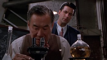 Movie still from “The Shadow” (1994), directed by Russell Mulcahy – A man looking through a microscope while another man looks on; Close Up shot, High angle