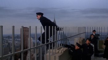 Movie still from “The Shadow” (1994), directed by Russell Mulcahy – A man in a black suit is jumping over a railing on the side of a building; Wide shot, Overhead angle