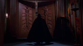 Movie still from “The Shadow” (1994), directed by Russell Mulcahy – A man in a black cape and hat standing in front of two doors; Wide shot, Low angle