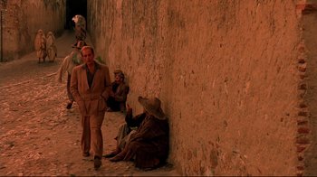 Movie still from “The Sheltering Sky” (1990), directed by Bernardo Bertolucci – A group of people sitting next to each other on a sidewalk; Wide shot, Low angle