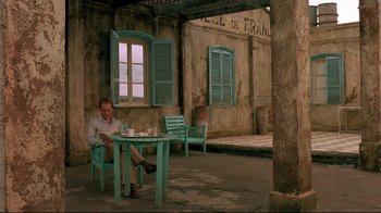 Movie still from “The Sheltering Sky” (1990), directed by Bernardo Bertolucci – A man sitting at a table in front of a building; Wide shot, High angle