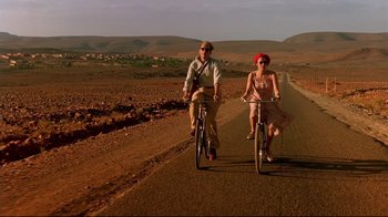 Movie still from “The Sheltering Sky” (1990), directed by Bernardo Bertolucci – A man and a woman riding bikes down a dirt road; Wide shot, Low angle