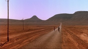 Movie still from “The Sheltering Sky” (1990), directed by Bernardo Bertolucci – Two people riding motorcycles down a dirt road in the middle of the desert; Extreme Wide shot, High angle