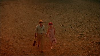 Movie still from “The Sheltering Sky” (1990), directed by Bernardo Bertolucci – A man and a woman holding hands while walking across a field; Wide shot, High angle
