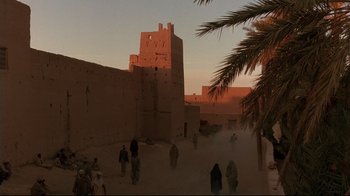 Movie still from “The Sheltering Sky” (1990), directed by Bernardo Bertolucci – A group of people walking in the sand near a building; Extreme Wide shot, Low angle
