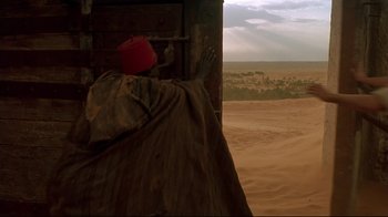 Movie still from “The Sheltering Sky” (1990), directed by Bernardo Bertolucci – A man in a red hat is standing in front of an open door in the desert; Wide shot, Over the shoulder angle