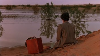 Movie still from “The Sheltering Sky” (1990), directed by Bernardo Bertolucci – A person sitting on a beach with a suitcase; Wide shot, High angle