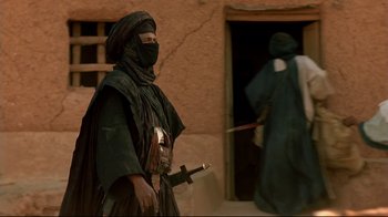 Movie still from “The Sheltering Sky” (1990), directed by Bernardo Bertolucci – A man in a burka holding a sword in front of a building; Medium shot, Over the shoulder angle