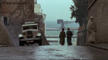 Movie still from “The Sheltering Sky” (1990), directed by Bernardo Bertolucci – Two men are walking down the street near an old car; Wide shot, High angle