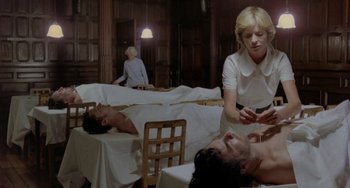 Movie still from “The Shout” (1978), directed by Jerzy Skolimowski – Two men laying on a bed in a room with a woman sitting at the table; Medium shot, High angle